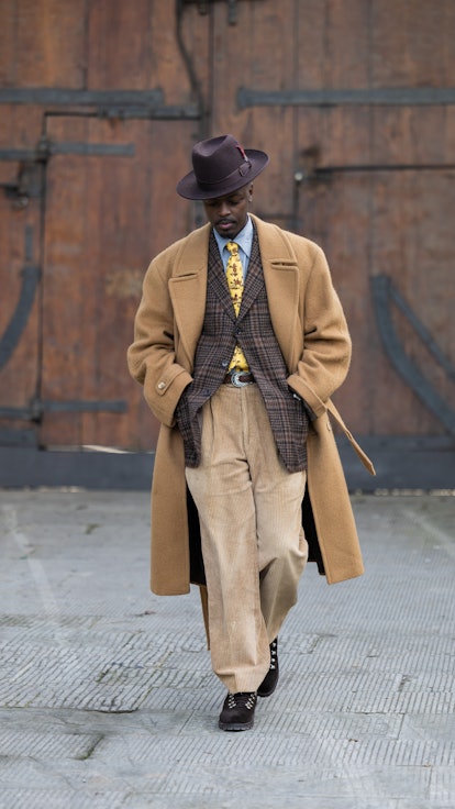A guest wears beige double breasted coat, brown hat, checkered blazer, pants at Fortezza Da Basso on...
