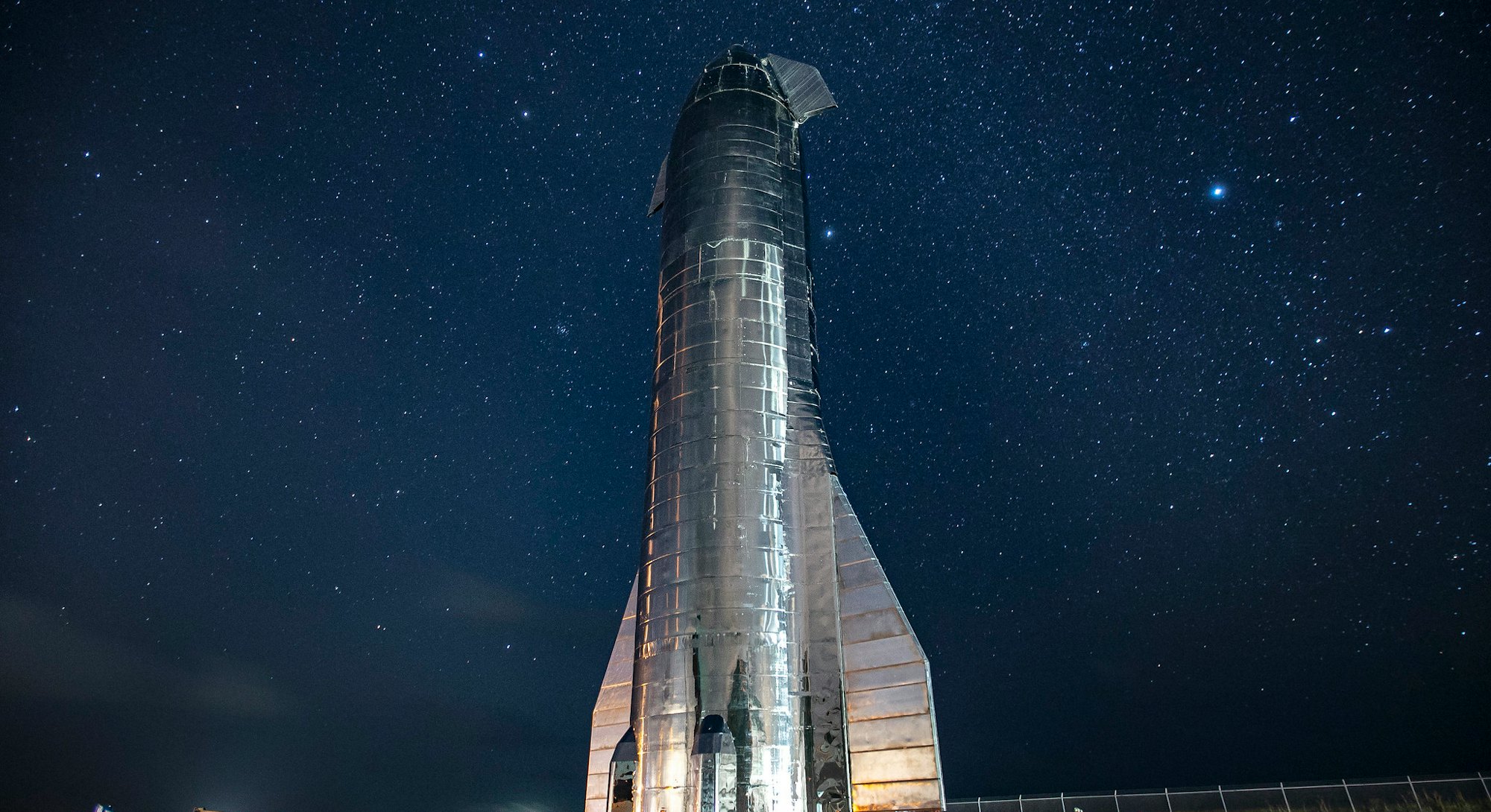 SpaceX's giant Starship launch vehicle towers over the land with glowing stars in the background aga...