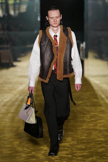 a model on a runway wearing a shearling vest and carrying three shopping-bag shaped leather bags