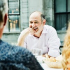 A man with a bald spot outdoors with friends, sitting at a table.