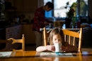 一个孩子在餐桌上用马克杯喝水，而她的父亲在厨房里。