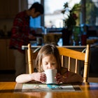 A child drinking from a mug at the dinner table while her father is in the kitchen.