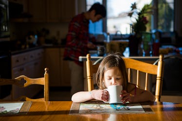 A child drinking from a mug at the dinner table while her father is in the kitchen.