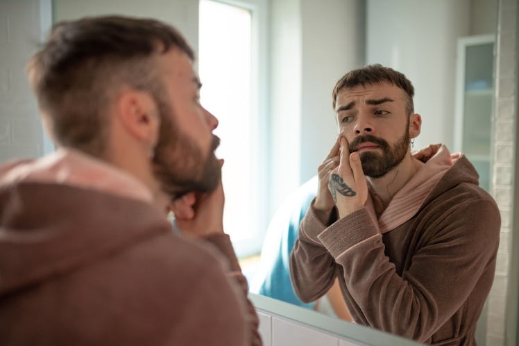 A man with a pimple picking at his face in the bathroom mirror.