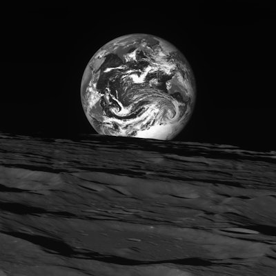 The Earth, appearing as a black and white marble with streaks of clouds across its atmosphere, peeks...