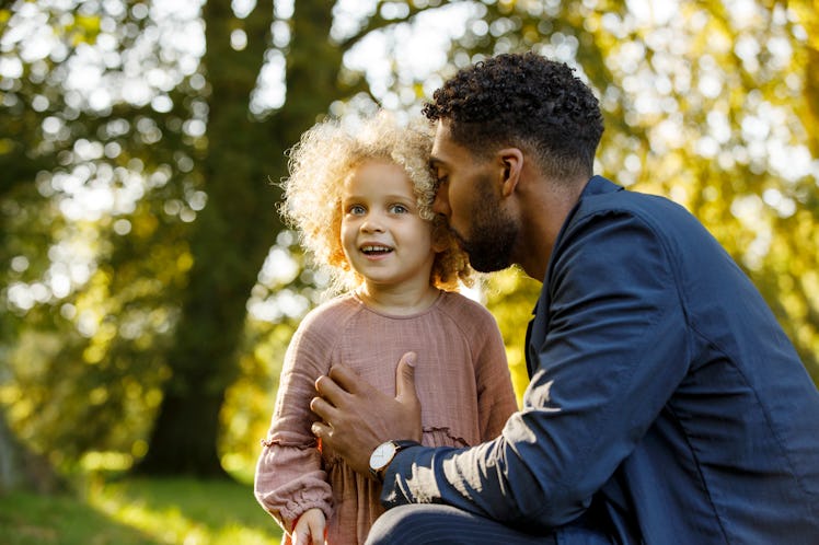 Middle aged father whispering something into delighted child's ear as they stand in tree-filtered su...