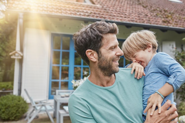A bearded dad smiles in the sunlight while holding his giggling young son