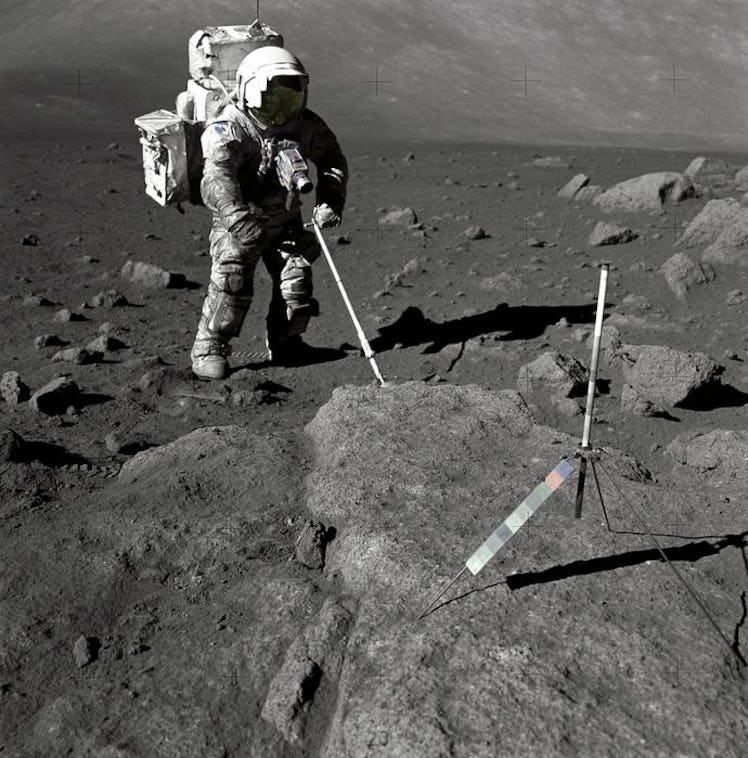 An Apollo astronaut stands on a gravely surface, prodding a boulder with a long stick. The astronaut...