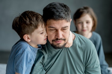 A dad looks uncomfortable as his son gives him a side hug, with his daughter in the background.