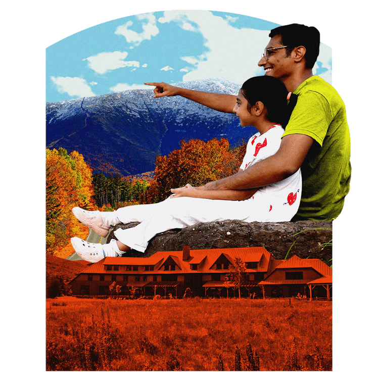 A parent sits with his child, pointing off in the distance. Behind them is fall foliage and some mou...