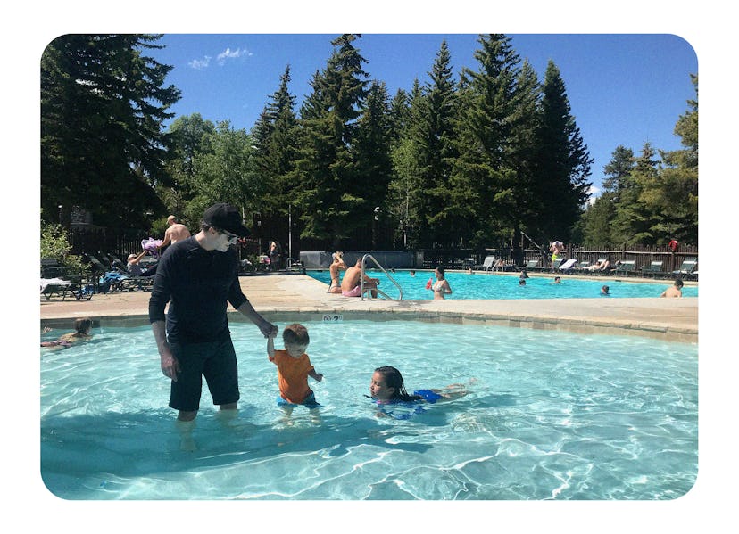 A father holds his son's hand in a pool, and his daughter swims next to them.