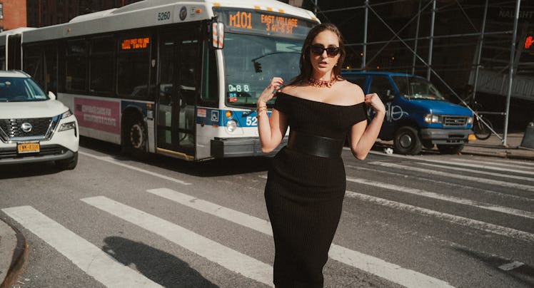 The comedian Catherine Cohen standing on a New York City crosswalk in a form-fitting black dress.