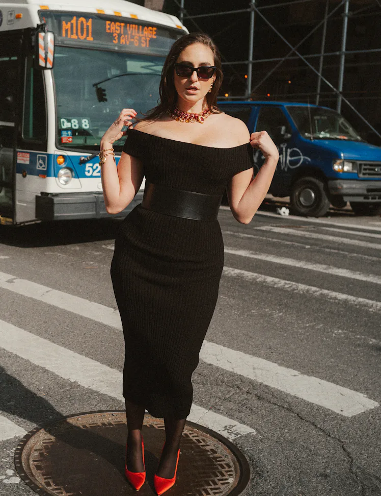 The comedian Catherine Cohen standing on a New York City crosswalk in a form-fitting black dress.