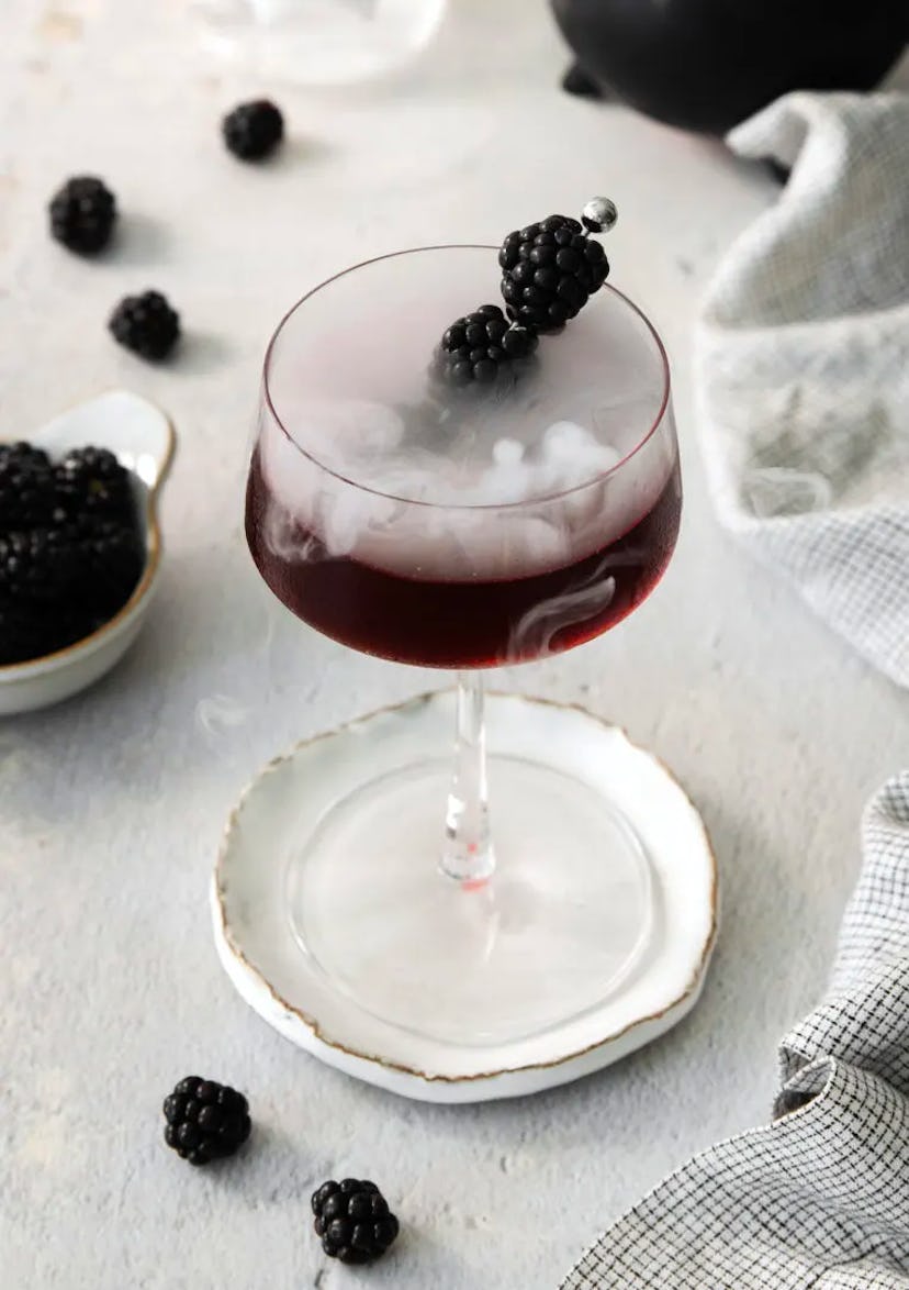 Halloween cocktail called a Black Widow, a dark drink garnished with blackberries in a coupe glass
