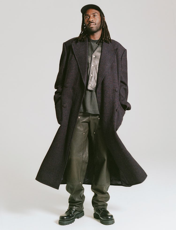 Devonté Hynes wears a charcoal wool trench coat, green leather pants, combat boots, leather vest and...