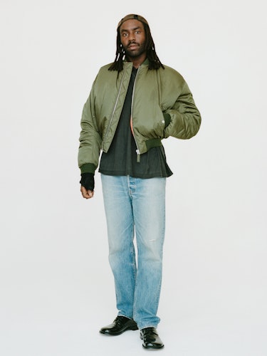 Devonté Hynes wears a green bomber jacket, denim jeans and shoes.