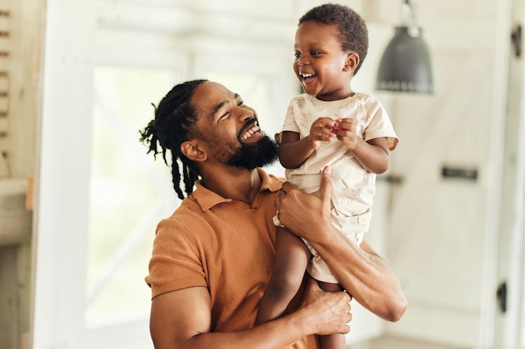 A dad holding his smiling toddler in their home.