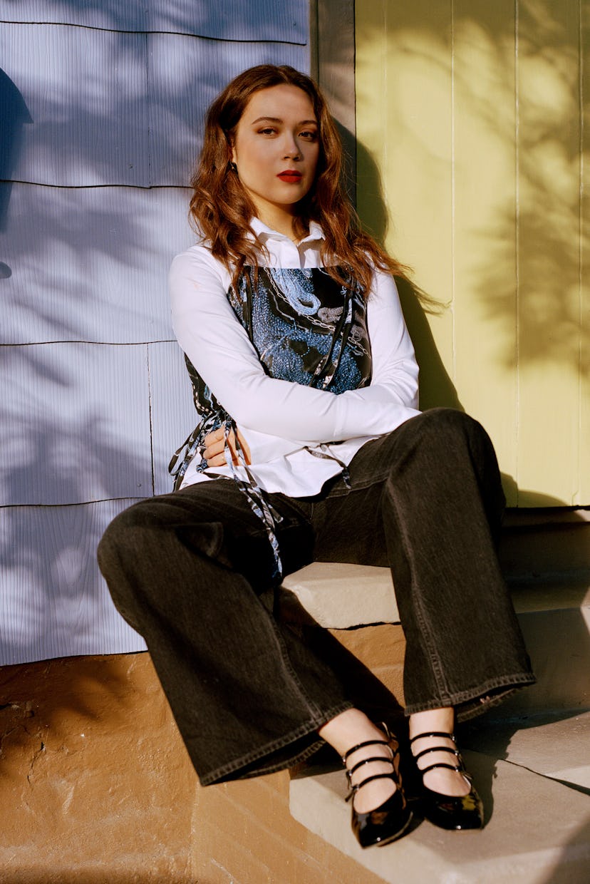 A young woman sits on steps, wearing denim overalls, a white blouse, and black sandals, bathed in su...