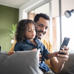 A dad sitting on a couch with his child in his lap, using a cell phone.