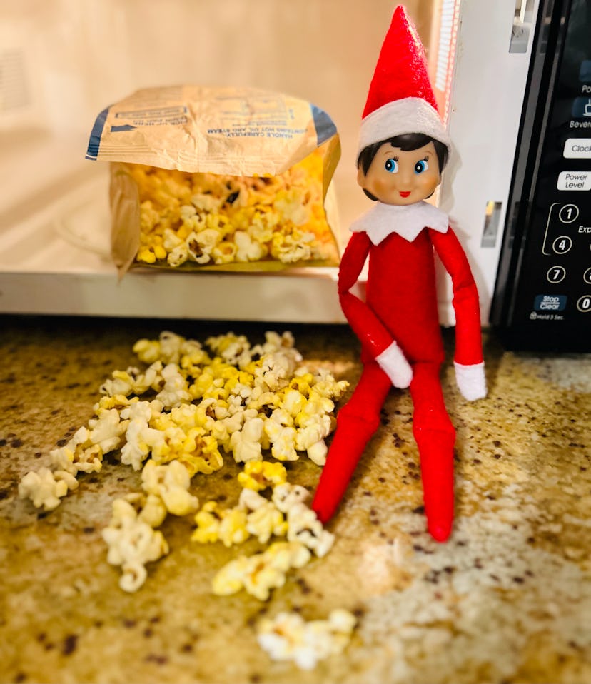 elf on the shelf sitting next to an open microwave with an open bag of popcorn in it