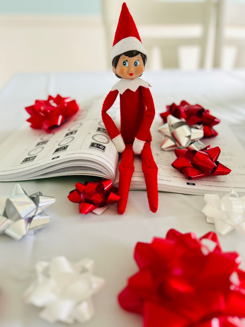 elf on the shelf sitting on top of an open homework book with ribbons around him