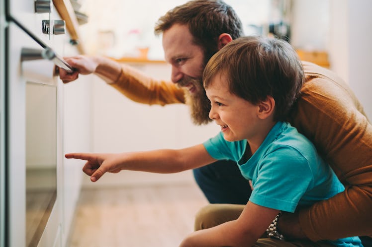 A dad and son looking at the food that they just put in the oven through the oven door, smiling.