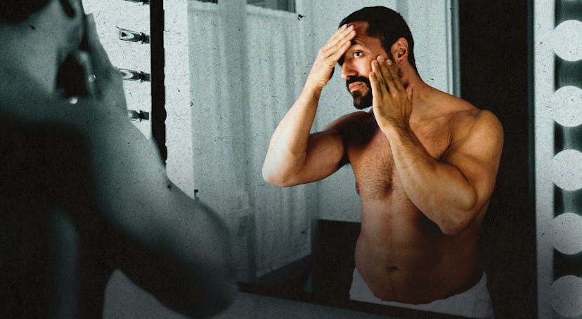 A shirtless man looking in the bathroom mirror, rubbing his face.