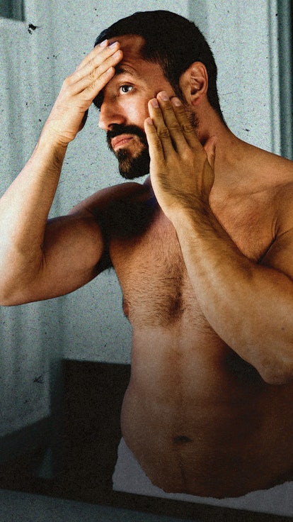 A shirtless man looking in the bathroom mirror, rubbing his face.