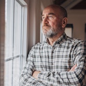 Man in checkered shirt looking worried as he stares out his window with his arms crossed