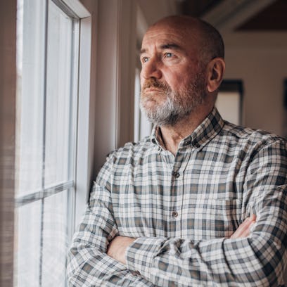 Man in checkered shirt looking worried as he stares out his window with his arms crossed