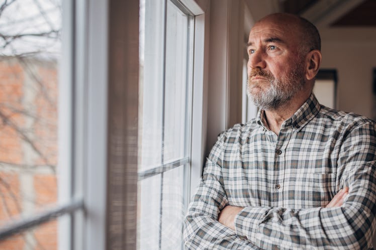 Man in checkered shirt looking worried as he stares out his window with his arms crossed