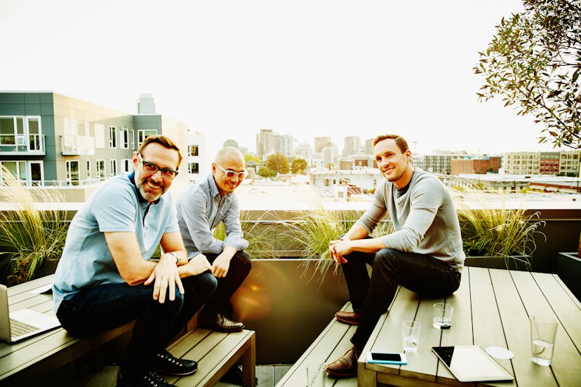 Three middled aged men who are friends sitting on a rooftop and having a conversation in the afterno...