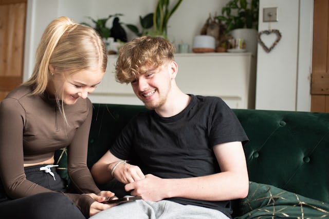 A young teen couple sits on the couch talking and playing on their phones.