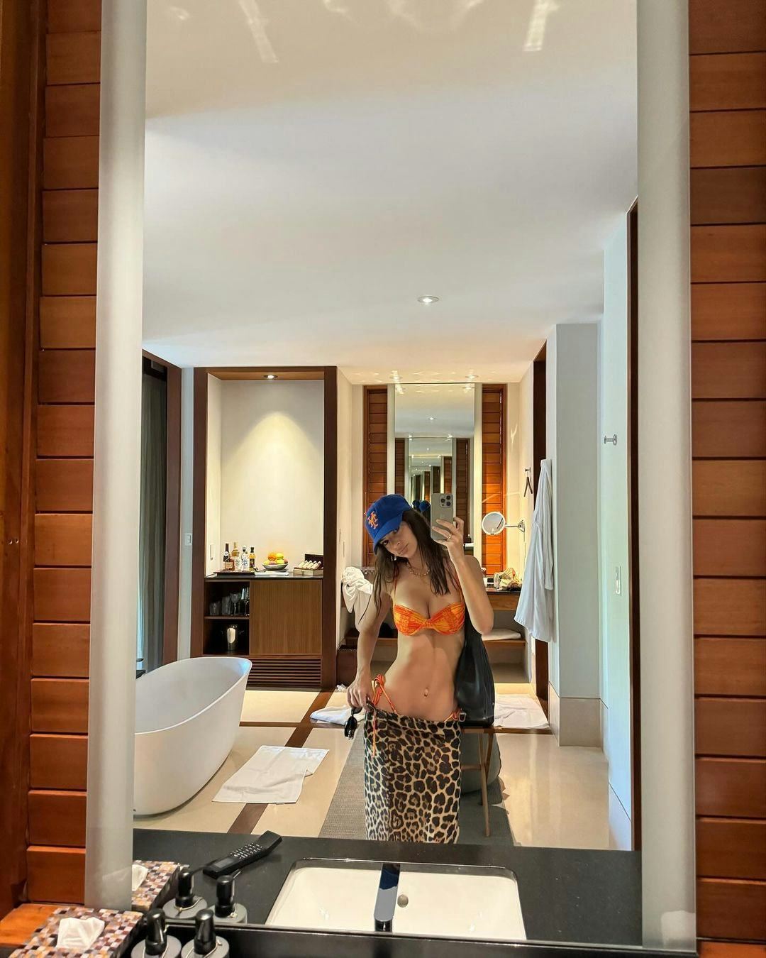 Emily Ratajkowski wears an orange bikini and baseball cap.