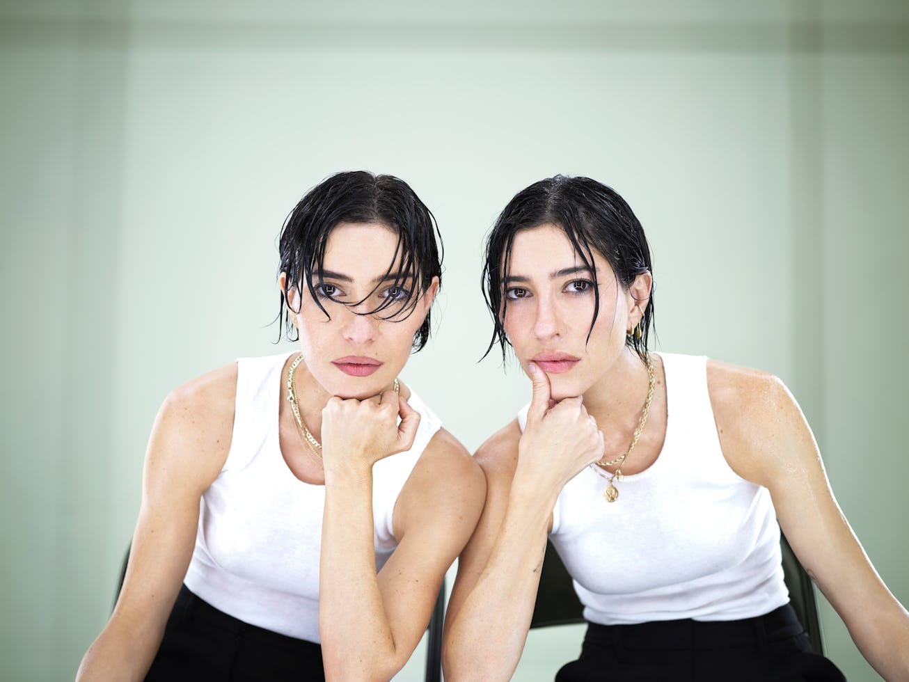 Two women with wet hair and white tank tops sitting and leaning forward, resting their chins on thei...