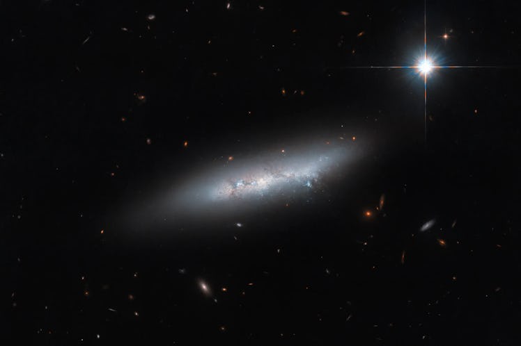 An irregular galaxy, a narrow streak of stars crossed by faint dust lanes. It is surrounded by a bri...