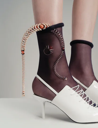 A model's feet wearing white heeled shoes and sheer black socks with shimmering jewelry visible thro...
