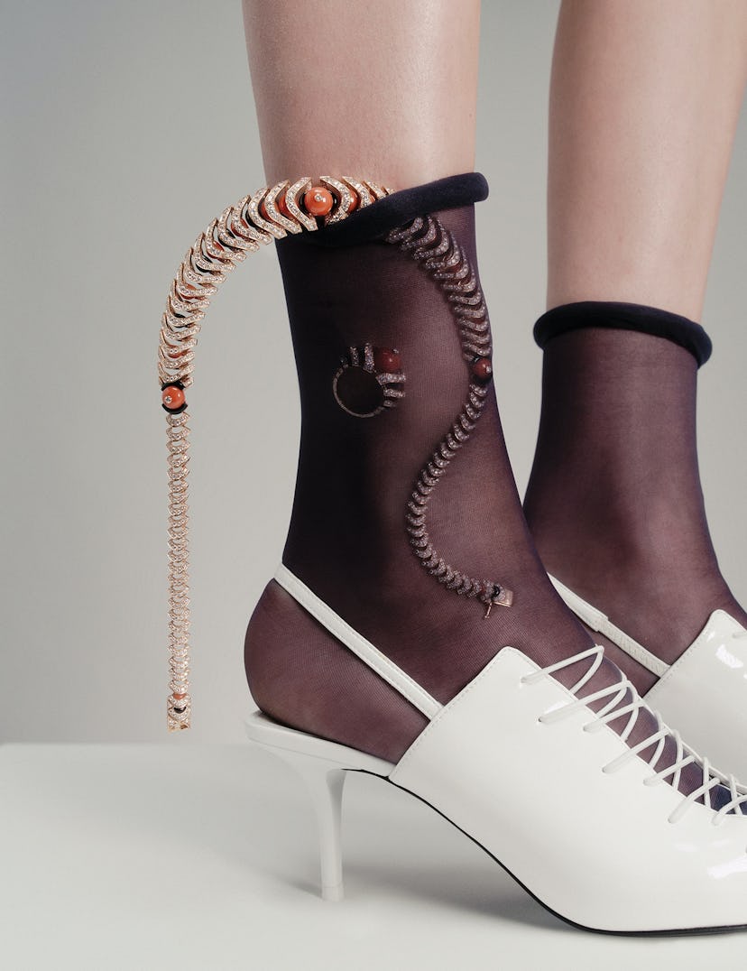 A model's feet wearing white heeled shoes and sheer black socks with shimmering jewelry visible thro...