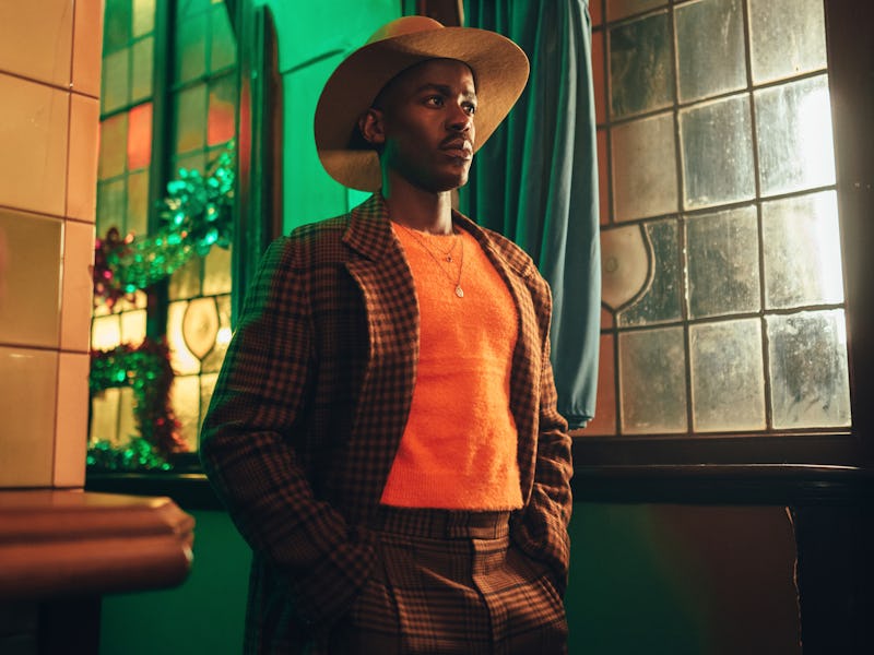 Man in stylish outfit with a wide-brimmed hat stands confidently indoors by a window with colorful n...