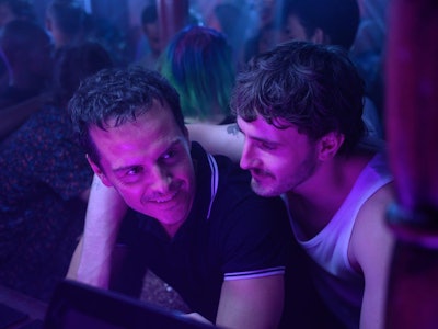 Two men smiling and leaning close to each other in a club with colorful lighting and people in the b...