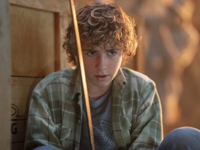 Young individual with curly hair and a plaid shirt looks concerned while hiding behind a wooden stru...