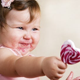A cute toddler with food on her face reaches out to give a lollipop.