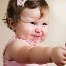 A cute toddler with food on her face reaches out to give a lollipop.