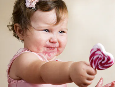 A cute toddler with food on her face reaches out to give a lollipop.