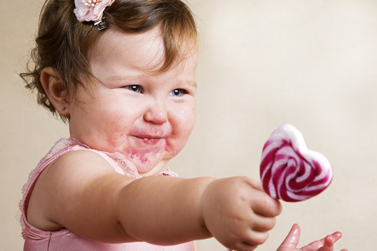 A cute toddler with food on her face reaches out to give a lollipop.