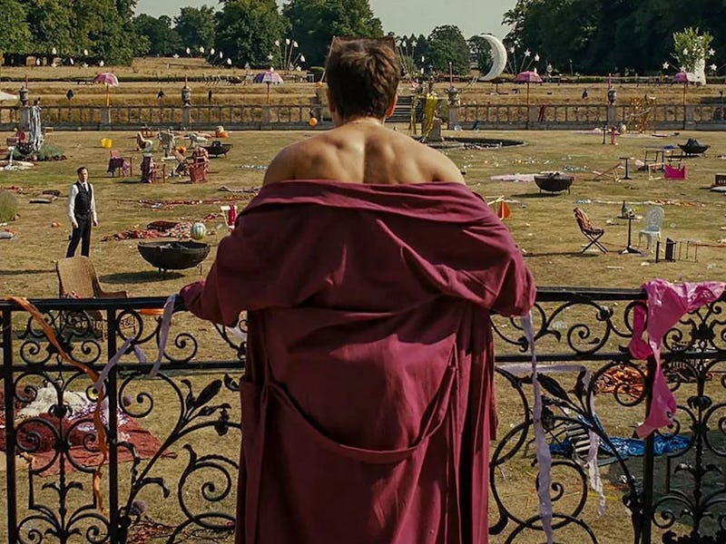 Man in a burgundy robe stands on a balcony overlooking a busy square with people and colorful decora...