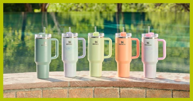 Five colorful insulated Stanley tumblers with lids and straws arranged on a poolside brick ledge wit...