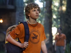 A young boy with curly hair, wearing an orange t-shirt and carrying a backpack, looks worried in a f...