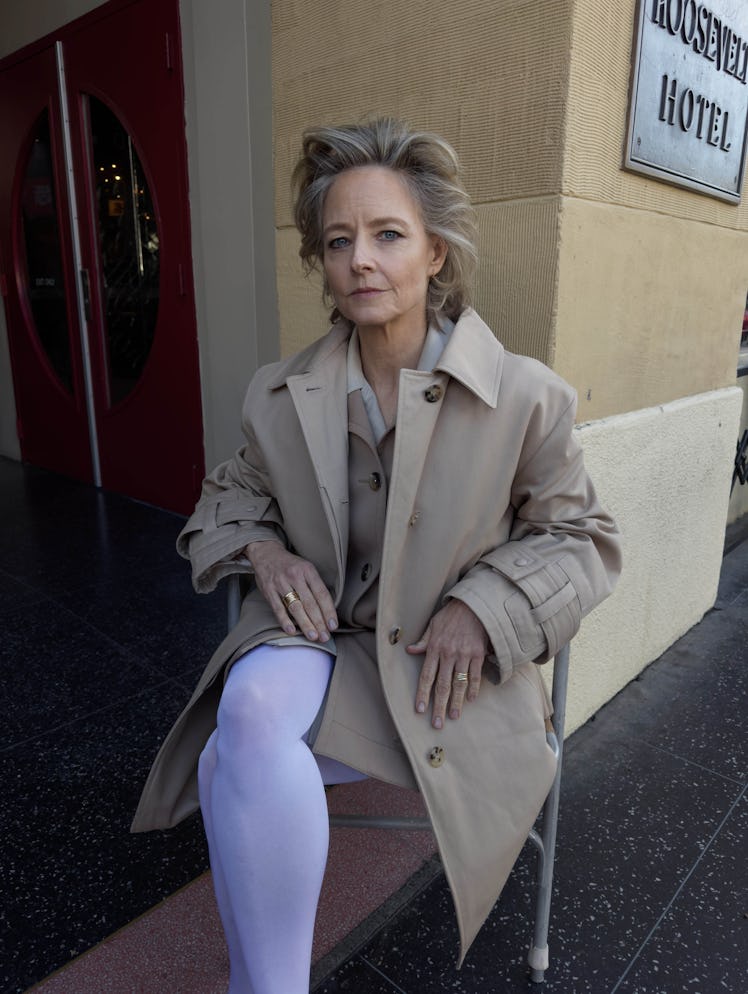 Jodie Foster wears a beige trench coat, beige vest, white tights, rings and earrings.