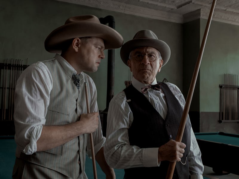 Two men in vintage attire with hats are holding a rifle, appearing to be in deep conversation in an ...